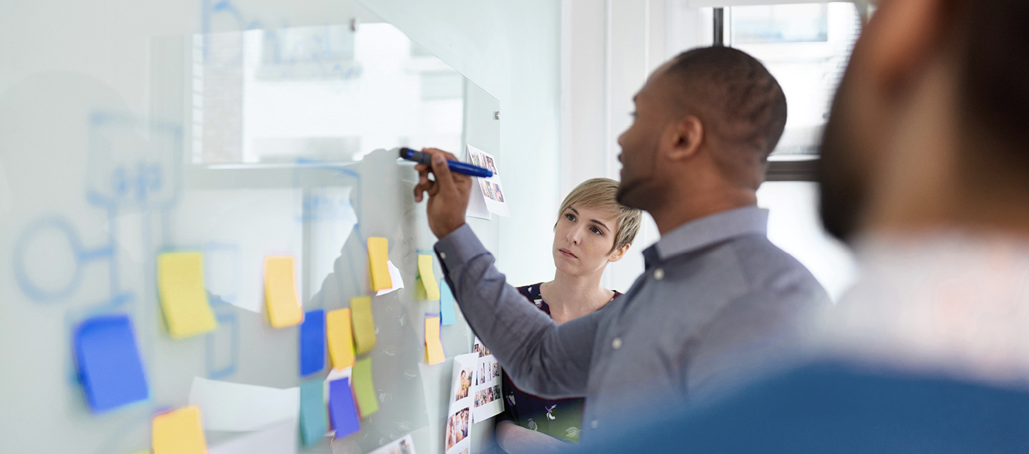 Ein Mann schreibt etwas auf ein mit Post-its beklebtes Whiteboard. Links und rechts von ihm stehen zwei Personen und schauen zu.