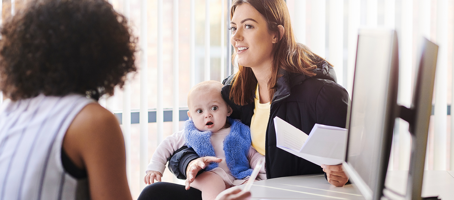 Eine Frau mit Baby auf dem Schoß sitzt mit einer weiteren Frau an einem Tisch. Sie unterhalten sich.