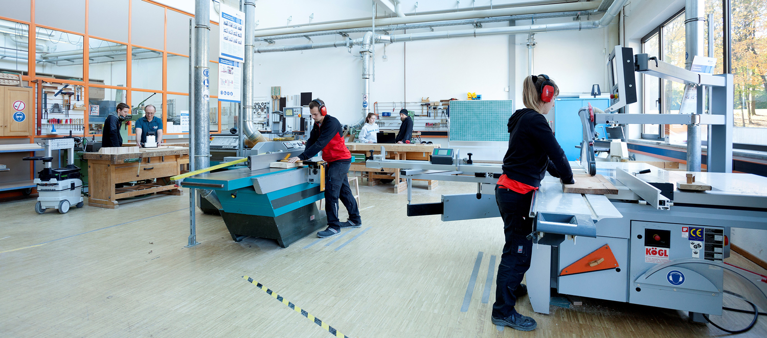 Mehrere Personen stehen in einer Werkstatt an Maschinen und Tischlerbänken.