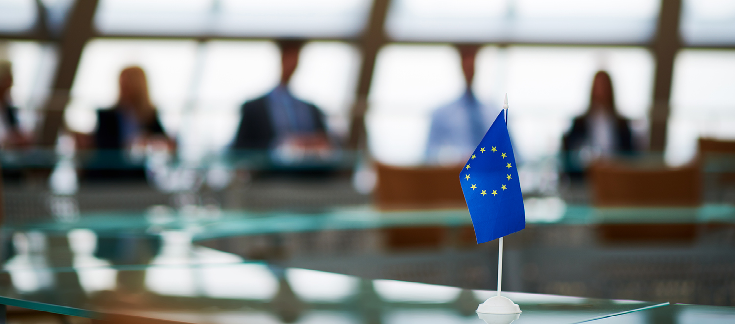 Ein Seminarraum. Im Hintergrund sitzen fünf Personen. Im Vordergrund ist eine kleine EU-Flagge auf dem Tisch zu sehen.