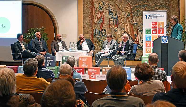 Sechs Männer sitzen auf einem Podium. Einer spricht in ein Mikrofon. Zuschauende sitzen in den Reihen vor ihnen.