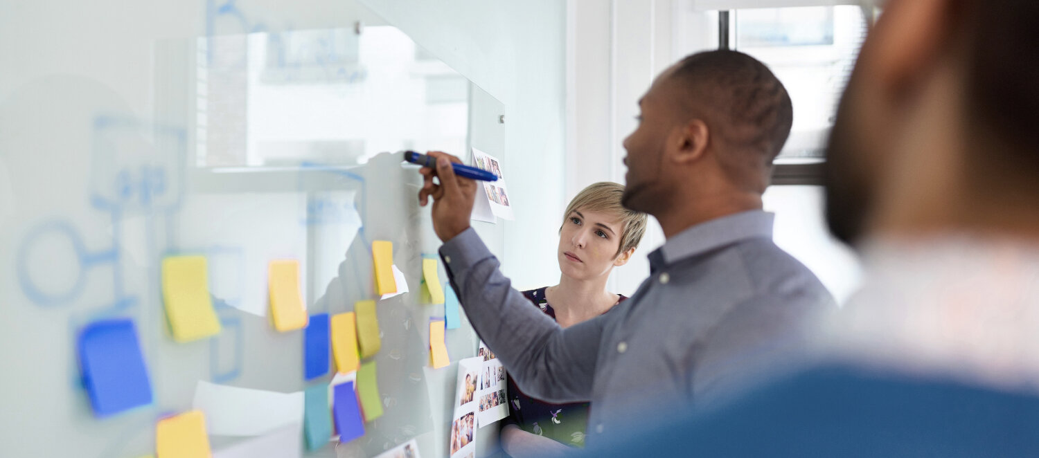 Ein Mann schreibt etwas auf ein mit Post-its beklebtes Whiteboard. Links und rechts von ihm stehen zwei Personen und schauen zu.